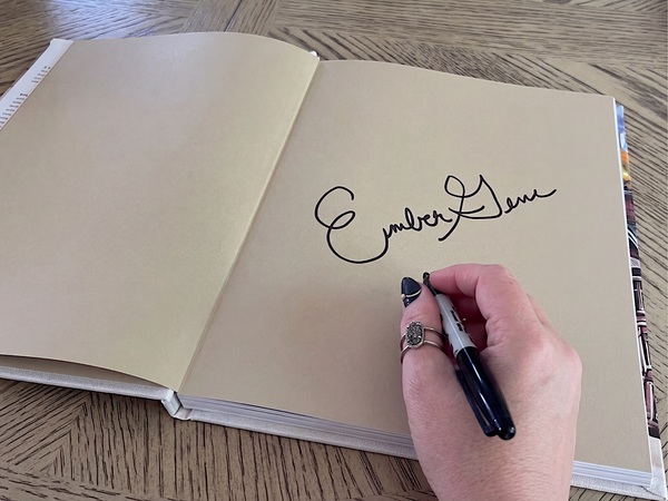 Close-up of author Ember Gene signing a copy of the children’s book Old Clothes with a black marker on the book’s front endpaper, shown on a wooden table