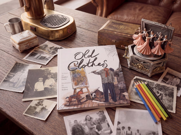 The children’s book Old Clothes displayed on a wooden table surrounded by vintage family photographs, colored pencils, antique keepsake boxes, and a dancing music box, creating a nostalgic old-fashioned atmosphere.
