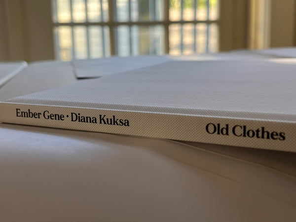 Close-up of the clothbound spine of the children’s book Old Clothes with the dust jacket removed, featuring the title, publisher logo, author Ember Gene, and illustrator Diana Kuksa printed in black on a textured white cover.